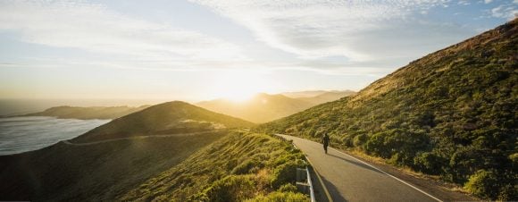 Person walking on winding mountain road looking into sun
