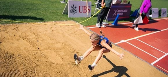 L’image montre l’épreuve du saut en longueur lors de l’UBS Kids Cup. Une fille effectue un saut en longueur dans le sable, dans un stade. À l’arrière-plan, on voit le logo UBS et les officiels (juges).