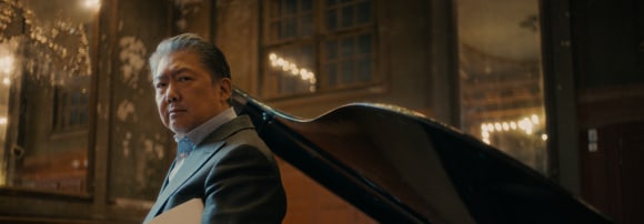 Der chinesische Dirigent klassischer Musik Long Yu steht mit einem Notenblatt in der Hand vor einem Klavier.