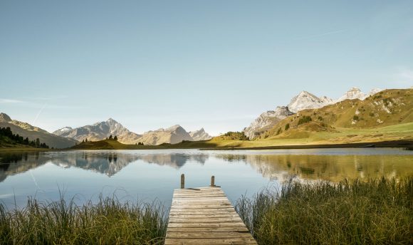 A wooden pier extends into a calm mountain lake surrounded by forested slopes and distant peaks.