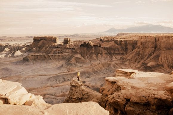 A long hiker in a yellow jacket stands on the edge of towering sandstone cliff