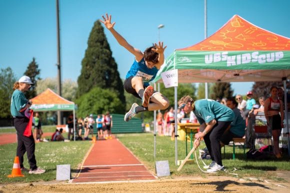 Ein dunkelhaariger Junge (ca. 12) springt beim Weitsprung auf dem Sportplatz, ein Fuss nach vorne, Arme oben. Zwei Erwachsene beobachten den Absprung. Im Hintergrund grüne Bäume, blauer Himmel, zwei UBS Kids Cup Pavillons (grün/orange) und wartende Kinder.