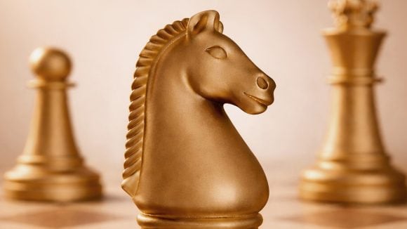 Close up view of a gold-hued chess board featuring the knight (horse) in the foreground and a pawn to the left and the queen to the right in the background.