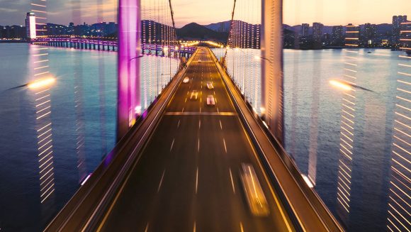 Aerial view of a suspension bridge at night with fast-moving cars and city lights in the distance.