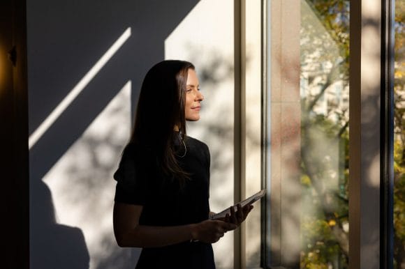 A women standing near a window with a book in her hand and the sunlight is reflecting on her