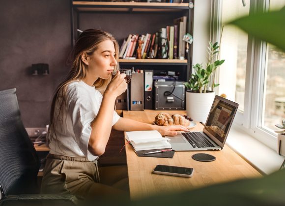Junge Frau am Schreibtisch sitzt teetrinkend vorm Laptop und schaut zum Fenster hinaus.