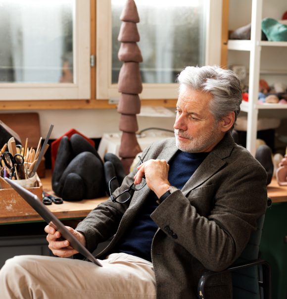 Ein Mann mit grauen Haaren und Bart sitzt auf einem Stuhl in einem Atelier und begutachtet ein Dokument, während er seine Brille in der linken Hand hält.