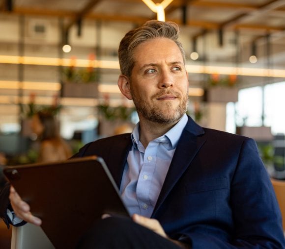 Blonder Mann mittleren Alters im Anzug sitzt mit einem Tablet in der Hand auf einem Stuhl und blickt in die Ferne.