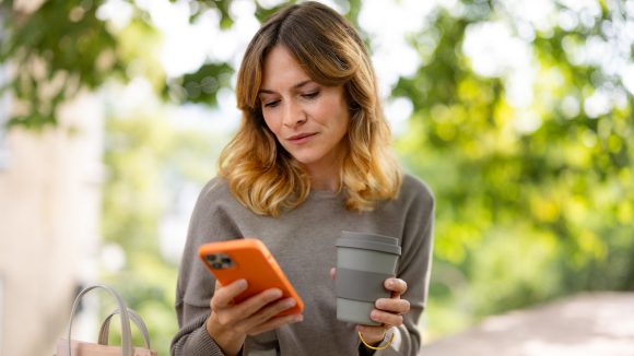 Eine Frau hält ihren Kaffee in der einen Hand und schaut auf ihr Smartphone in der anderen Hand.
