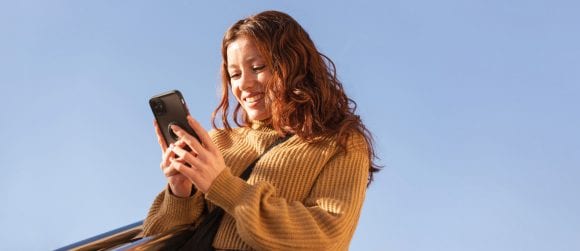 Red-haired woman in ochre sweater smiling at a smartphone