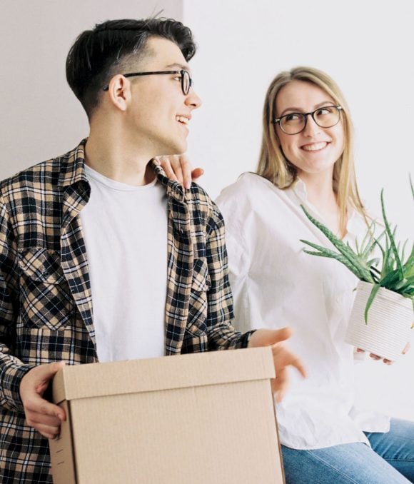 A laughing young couple is holding a moving box and a potted plant.