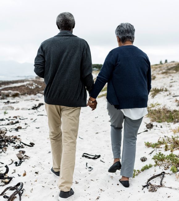 Older couple walking hand in hand on the beach (seen from behind).