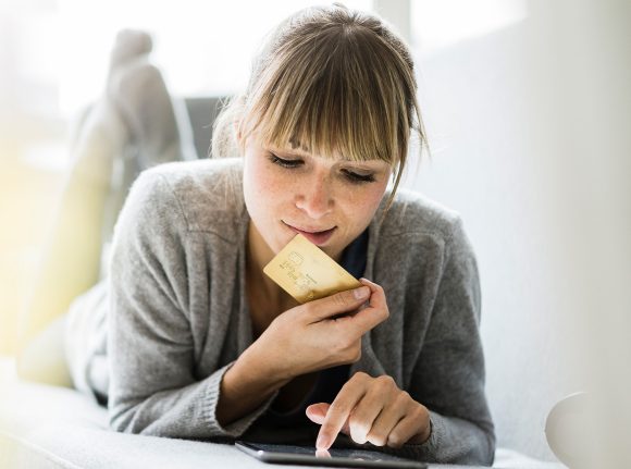 A young woman lying on the sofa using her tablet while holding her UBS Credit Card in her hand.