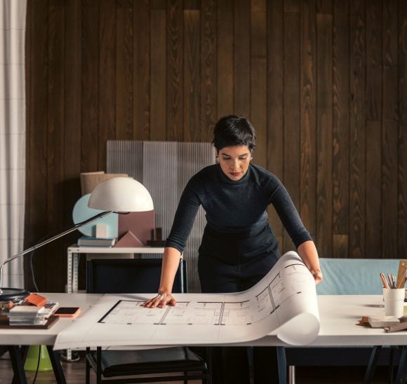 Une femme aux cheveux noirs et courts est assise à une table et consulte un grand plan déroulé qui ressemble à un plan de construction.