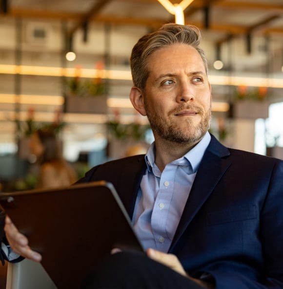 Un homme blond d’âge moyen en costume est assis sur une chaise avec une tablette à la main et regarde au loin.