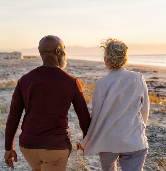 An elderly couple is walking hand in hand on a beach.