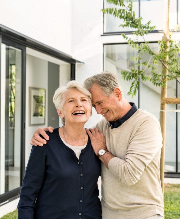 An older couple is standing in a garden in front of a house, laughing.