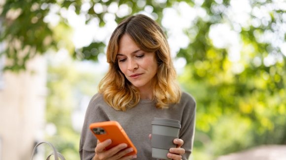 Eine Frau hält ihren Kaffee in der einen Hand und schaut auf ihr Smartphone in der anderen Hand.