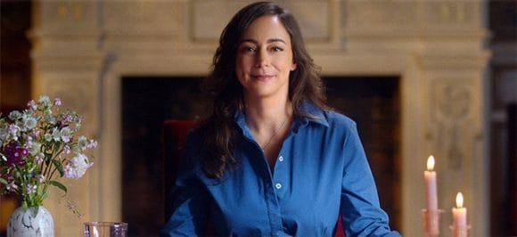 Eine Frau, die an einem Tisch sitzt und in die Kamera schaut.