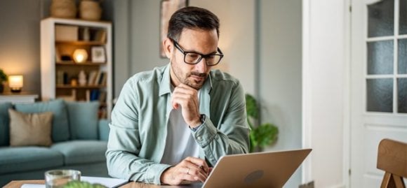 In diesem Bild sieht man einen jüngeren Mann im Wohnzimmer an einem Tisch sitzend und schaut konzentriert in einen Laptop.