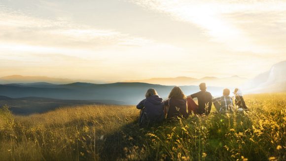A group of four individuals and their dog together on a hillside gazing into a sunset
