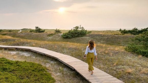 Woman with a hat walking barefoot along a boardwalk across low dunes toward the ocean with sun shining from beneath clouds.