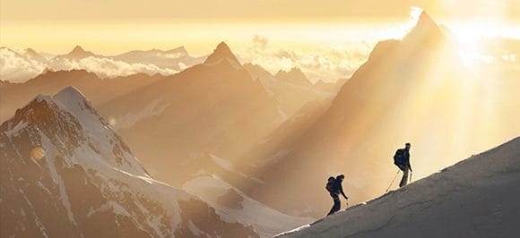 The picture shows two ski tourers climbing the side of a mountain. They are illuminated by the morning sun.