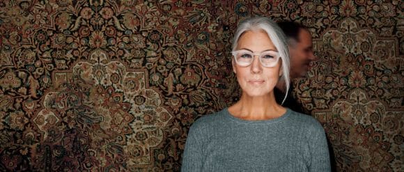 A lady with grey hair and silver glasses stands in front of a patterned carpet.