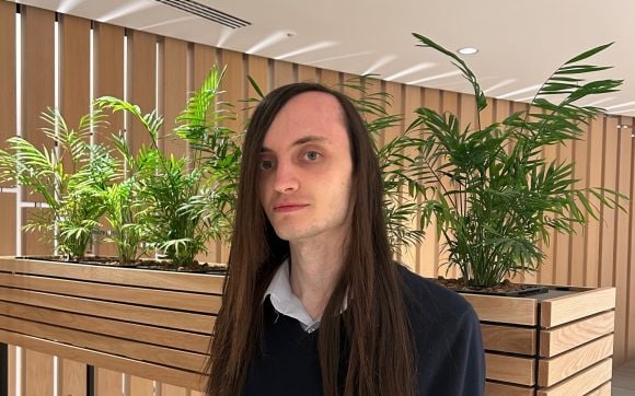 The image features a photo of Lewis standing in the UBS office area, dressed in a navy blue sweater with a visible collar of a white shirt. Lewis has long dark hair reaching past his shoulders. There is a wall of wooden panels and pots with green plants in the background.