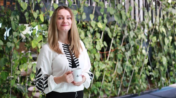 The image features a photo of Olga standing in the UBS office area, dressed in a white shirt with black plant motif and black pants. Olga has long blond hair reaching past her shoulders. She smiles while holding a branded cup with UBS logo. There is a green plants wall in the background.