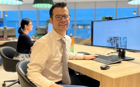 The image features a photo of Michael who seats at his desk and smiles to the camera with the office open space area in the background. Michael has short dark hair and glasses. He’s dressed in a white shirt with a gray tie and navy blue pants.