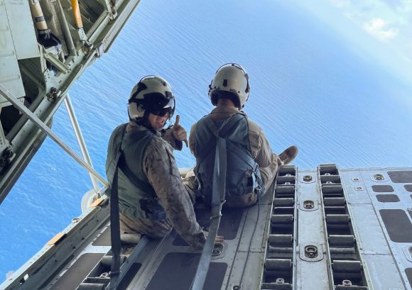 two men in an airplane. The one on the left is looking at the camera and giving a "hang loose" sign to the camera. the other is looking down at the water below them