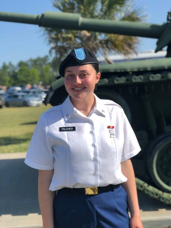 Photo of Skylar, who’s featured as a Veteran. She’s in uniform, standing in front of a military vehicle.