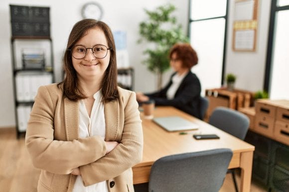 Portrait of a young woman with down syndrome smiling against the office area in the background.