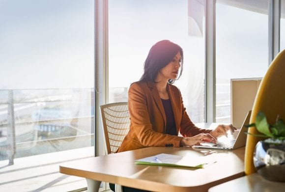 A woman in a sunny office writing on a laptop