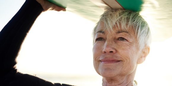 A woman with short hair and a wetsuit holding a surfboard on her head