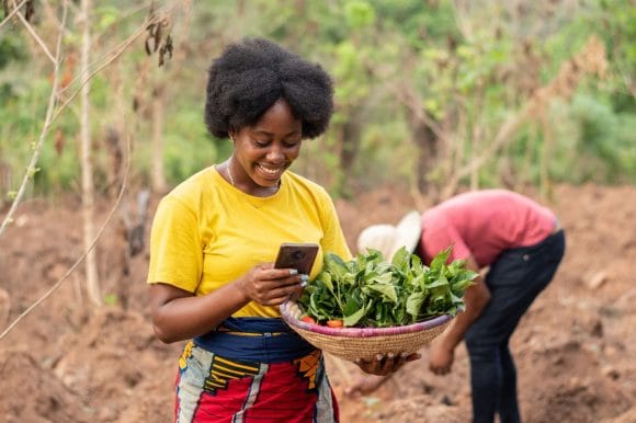 Smiling woman holding a basket of vegetables and using a smartphone in a rural setting