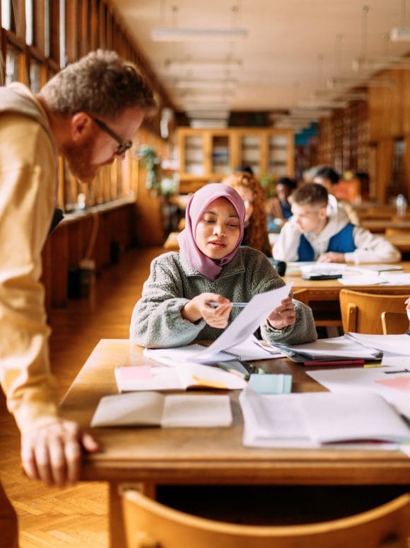 A young student in a library showing a paper to a teacher