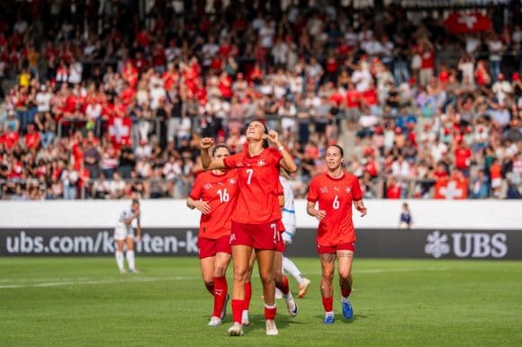Four members of a women’s football team are visible, showing strong emotion. Some members of the opposing team and a packed crowd of supporters in the stands can be seen in the background. The UBS logo is visible on the advertisement banner surrounding the pitch.
