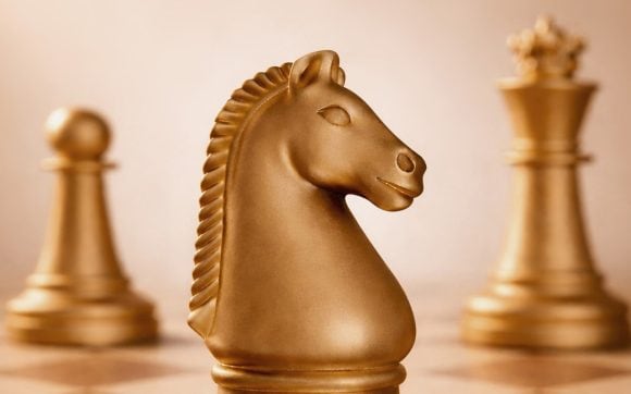 Close up view of a gold-hued chess board featuring the knight (horse) in the foreground and a pawn to the left and the queen to the right in the background.
