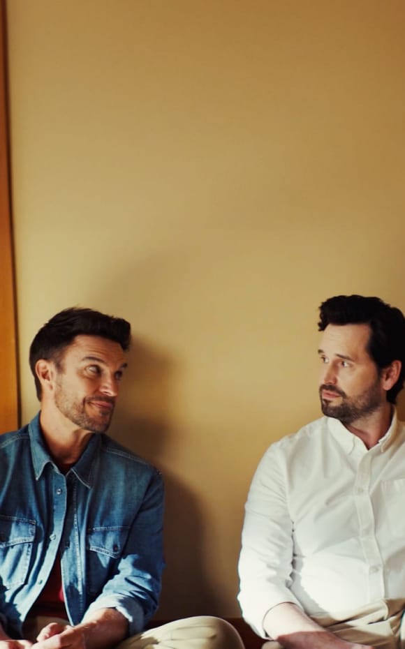 Two men sitting against a yellow wall having a conversation