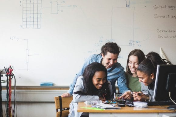 Smiling female teacher and high school teenage students preparing robot on desk in classroom