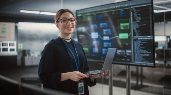 Portrait of a Diverse Female Wearing Glasses, Using Laptop Computer, Looking at Camera and Smiling. Information Technology Specialist, Software Engineer or Developer